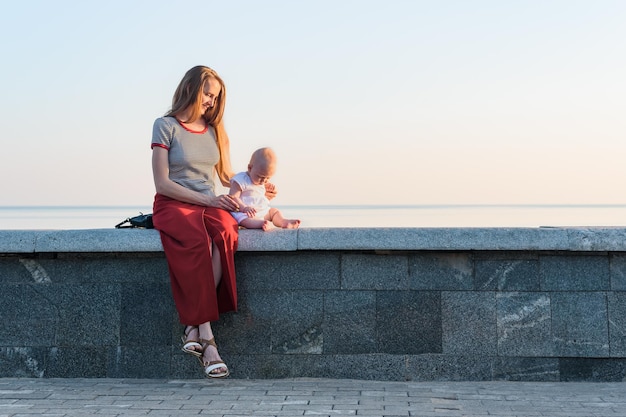 Young beautiful woman and toddler sitting on promenade on sea background Vacation with baby