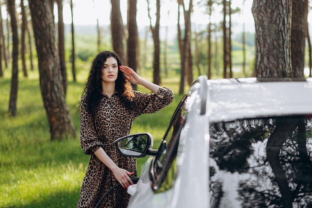 Young beautiful woman traveling by electric car having stop at charging station standing plugging cable browsing internet on smartphone smiling joyful while charing