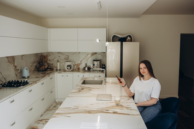 Young beautiful woman using mobile phone while healthy breakfast in light modern kitchen