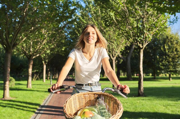 Young beautiful woman on vintage bike with basket rides on nature, sunny summer day alley in green park background, looking ahead