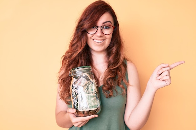 Photo young beautiful woman wearing glasses holding jar with savings smiling happy pointing with hand and finger to the side