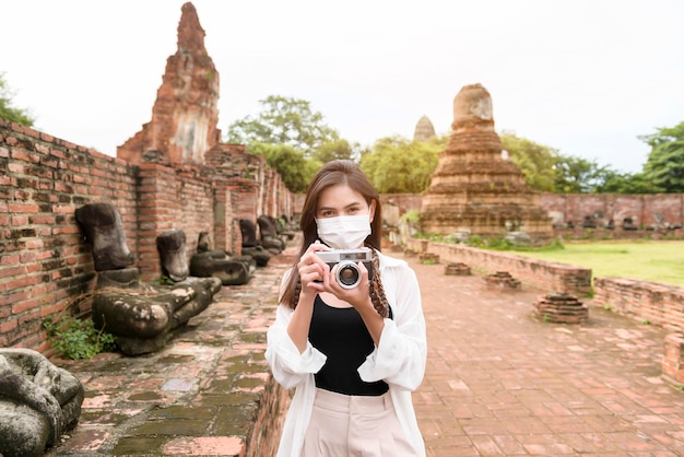 Young beautiful woman wearing protective mask traveling and taking photo at Thai historical Park