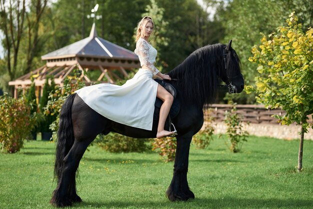 Young beautiful woman wearing a white dress riding horse, black horse, summer.