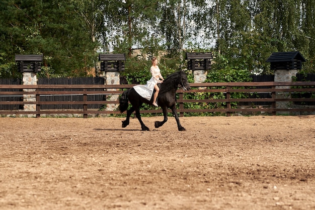 Giovane bella donna che indossa un abito bianco a cavallo, cavallo nero, estate.