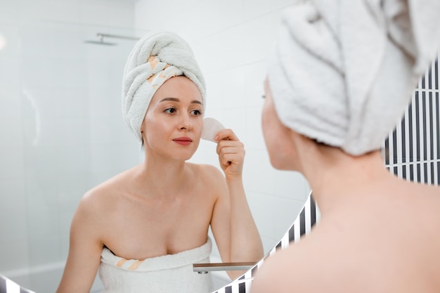 Young beautiful woman wearing white towel doing face massage with pink quartz scraper during beauty morning routine in bathroom Anti aging treatments and skincare concept