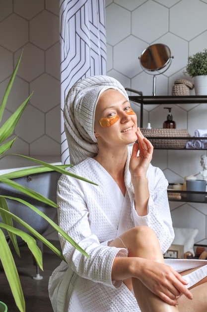 Young beautiful woman in a white coat applies golden collagen hydrogel patches under her eyes stands in the bathroom enjoys facial skin care treatments