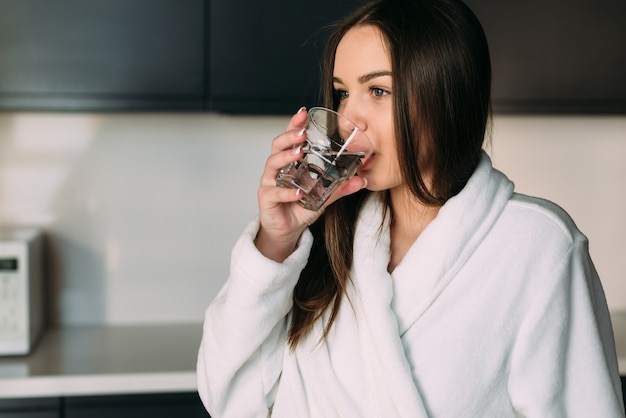 Young beautiful woman in a white coat in the kitchen drinks a glass of clean water.