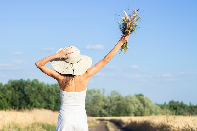 Young beautiful woman in a white dress and hat holds a bouquet with wildflowers on the road between the milky field. 