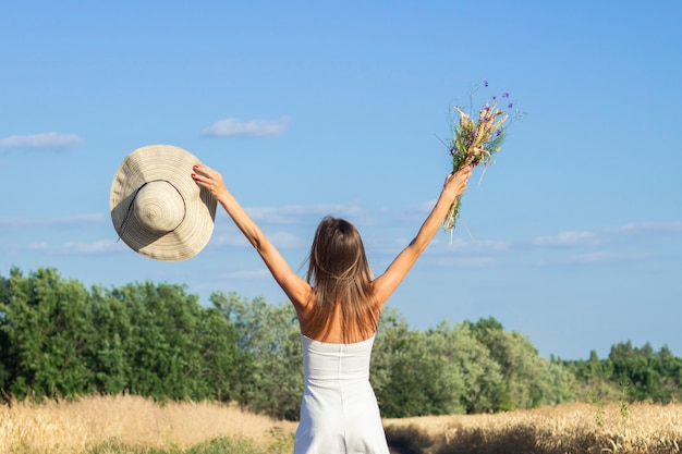 Young beautiful woman in a white dress and hat holds a bouquet with wildflowers on the road between the milky field. 