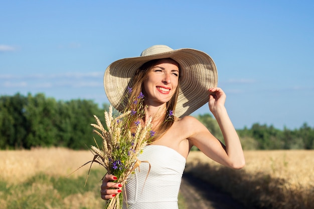 Young beautiful woman in a white dress and hat holds a bouquet with wildflowers on the road between the milky field. 