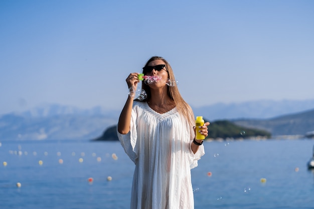 Young beautiful woman in white dress and sunglasses blowing soap bubbles on pier with sea view background. The concept of joy, ease and freedom during the vacation. The girl is enjoying the rest.
