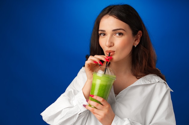 Young beautiful woman in white shirt drinking green smoothie against blue background