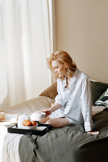 白いシャツを着た若い美しい女性がベッドに座って、朝の朝食を持っています。