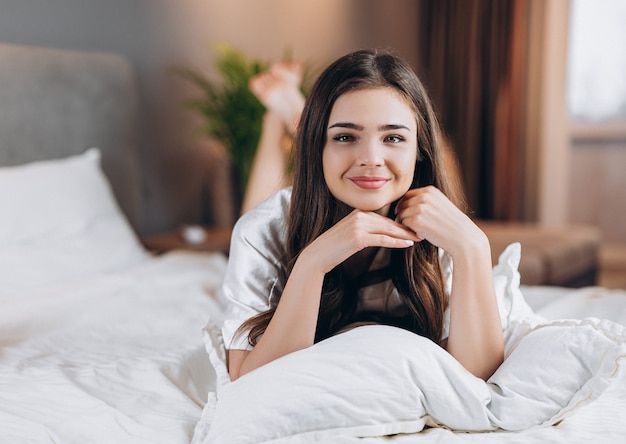 Young beautiful woman in white silk pajamas lies on the bed and smiles. Portrait of a beautiful female model lying on the bed.