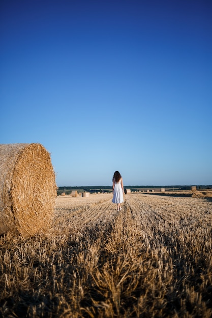 白い夏のドレスを着た若い美しい女性が、干し草の巨大な束のある刈り取られた麦畑に立って、自然を楽しんでいます。村の自然。セレクティブフォーカス