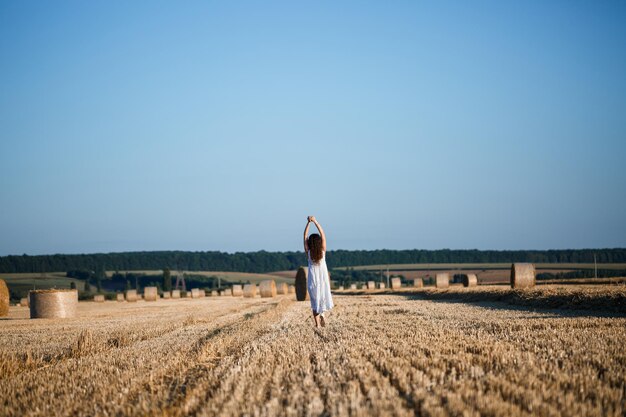 白い夏のドレスを着た若い美しい女性は、干し草の巨大な束のある刈り取られた麦畑に立って、自然を楽しんでいます。村の自然。セレクティブフォーカス