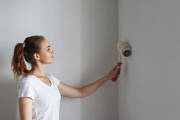 Young beautiful woman in a white T-shirt paints a wall with a roller in a new apartment during a renovation