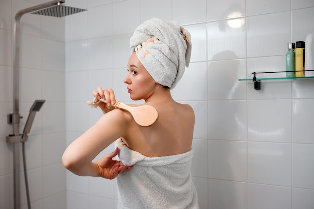 Young beautiful woman in white towel doing body lymphatic drainage massage with dry wooden brush with natural bristles in bathroom at home Anticellulite exercises
