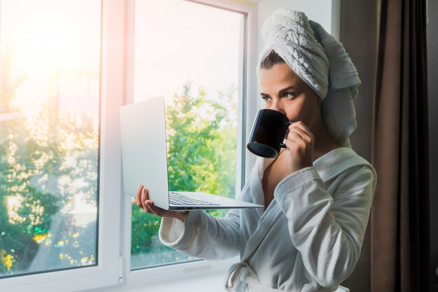 Giovane bella donna in asciugamano bianco e abito con un computer portatile in mano a bere il caffè vicino alla finestra