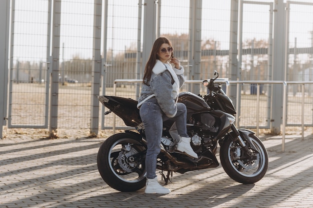 Young beautiful woman with black hair in denim pants and a jacket near a black sports motorcycle