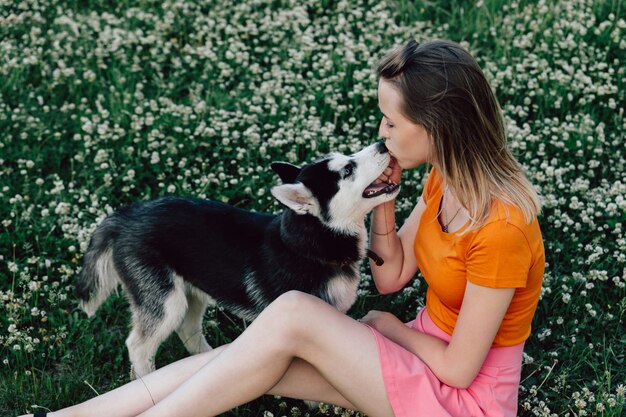 A young beautiful woman with blond hair is sitting in the meadow with her pet puppy husky and kissing him on the nose.