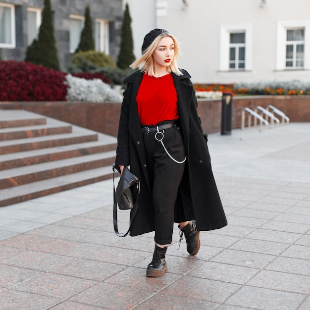 スタイリッシュなベレー帽の黒い秋のファッショナブルなアウターウェアの革のハンドバッグとブロンドの髪の若い美しい女性は、暖かい秋の日に街を歩きます