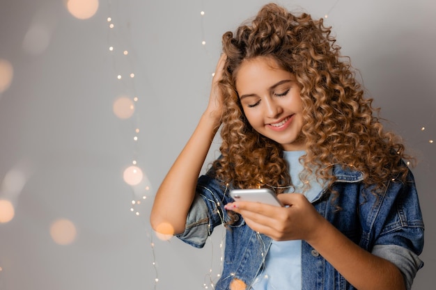 데님 재킷에 금발 곱슬머리를 한 젊고 아름다운 여성이 손에 스마트폰을 들고 있습니다. 소녀는 소셜 네트워크에서 온라인으로 의사 소통합니다. 블로거. 텍스트를 위한 공간입니다. 고품질 사진