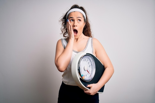 Young beautiful woman with curly hair holding weighing machine over white background afraid and shocked surprise and amazed expression with hands on face