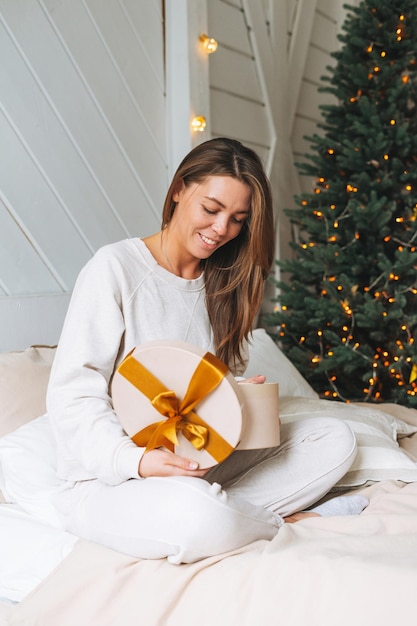 Young beautiful woman with dark long hair in cozy white pajamas with present gift box withgold ribbon sitting on bed in room with Christmas tree at the home Happy New Year