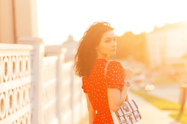 Young beautiful woman with a fashionable leather bag in a red summer dress are walking in the city at sunset