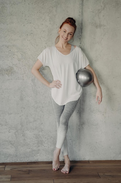 Молодая красивая женщина с фитболом позирует на бетонной стене студии после тренировки пилатесбарре, улыбаясь в камеру