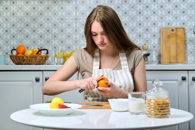 Giovane bella donna con frutta in cucina, donna seduta a tavola e pulizia arancione. food blogger femminile che cucina macedonia sulla macchina fotografica