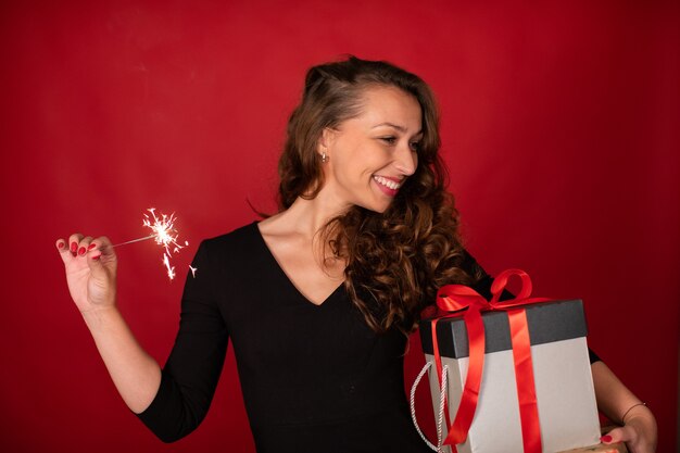 Young beautiful woman with gift box and burning sparkler smiling on red background. Christmas holiday party, New Year's Eve celebration, romantic gifting ideas, self-gifter concept. Copy space