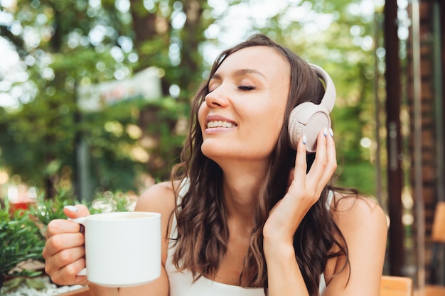 A young beautiful woman with headphones is sitting at a table in a summer cafe and drinking coffee or tea generation z