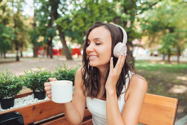 A young beautiful woman with headphones is sitting at a table in a summer cafe and drinking coffee or tea generation z