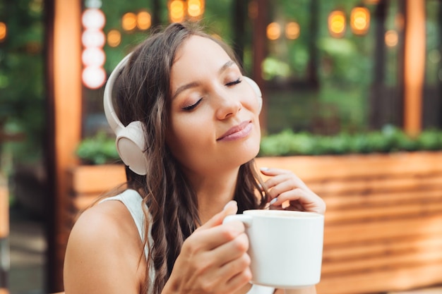 A young beautiful woman with headphones is sitting at a table in a summer cafe and drinking coffee or tea generation z