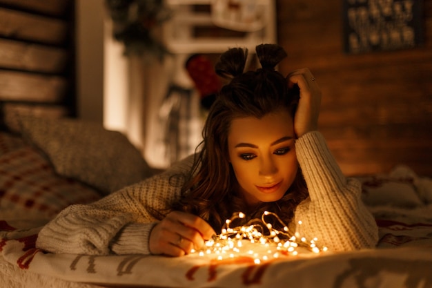 young beautiful woman with holiday lights in knitted retro sweater on the bed