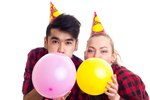 Young beautiful woman with long chestnut ponytail and young handsome man in red plaid shirts with celebrating hats blowing balloons on white background in studio