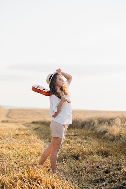 Young beautiful woman with long curly hair in shorts, a hat with a guitar poses in a wheat field in the summer at sunset