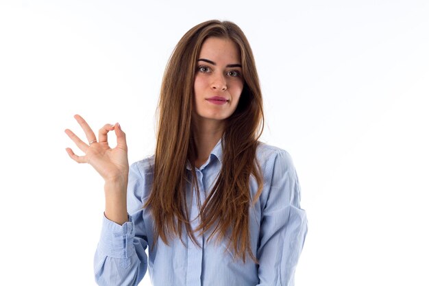 Young beautiful woman with long hair in blue shirt showing okay on white background in studio