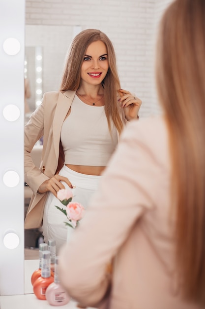 Young beautiful woman with long healthy blonde hair looking at the mirror in hairdressing salon