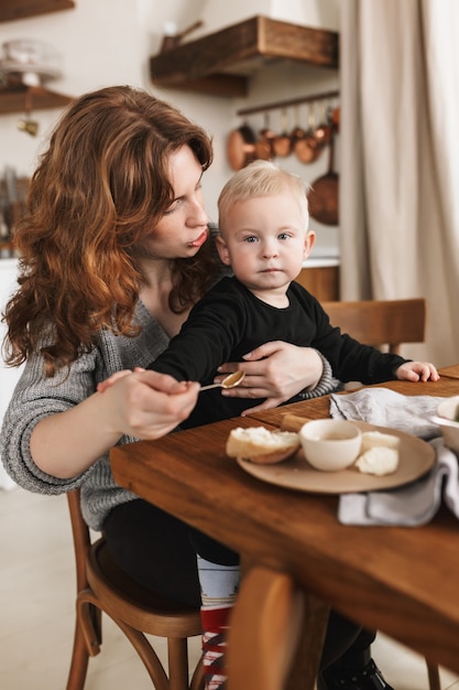 写真 夢のように彼女の幼い息子を養う食べ物と一緒にテーブルに座っているニットセーターの赤い髪の若い美しい女性