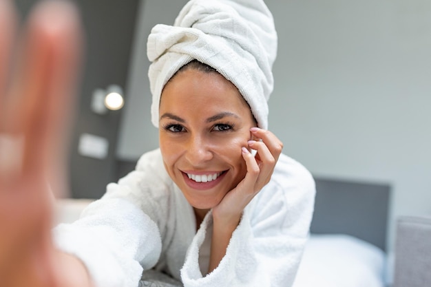 Foto giovane bella donna con un asciugamano su un letto in una stanza d'albergo vestita con un accappatoio bianco che si fa un selfie con il suo cellulare un leggero sorriso sul suo viso