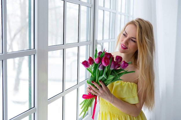 Young beautiful woman with tulip bunch in yellow dress looks at window 8 march international womens day