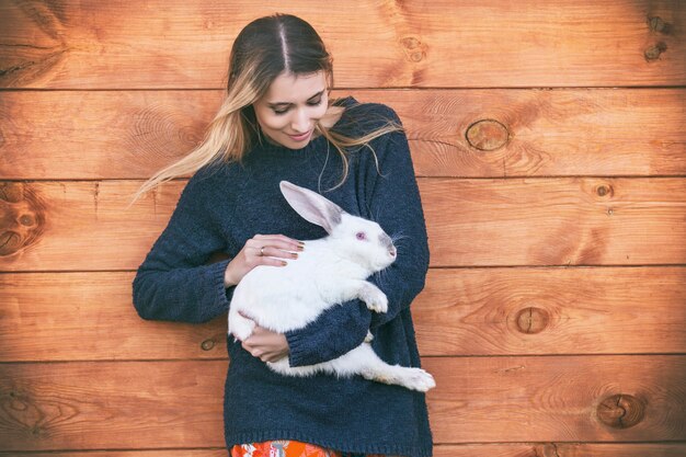 Young beautiful woman with a white rabbit in her arms next to a wooden house in the countryside