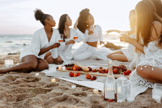 사진 해가 지는 해변에서 와인과 과일로 여름 피크닉을 하는 아름다운 젊은 여성들