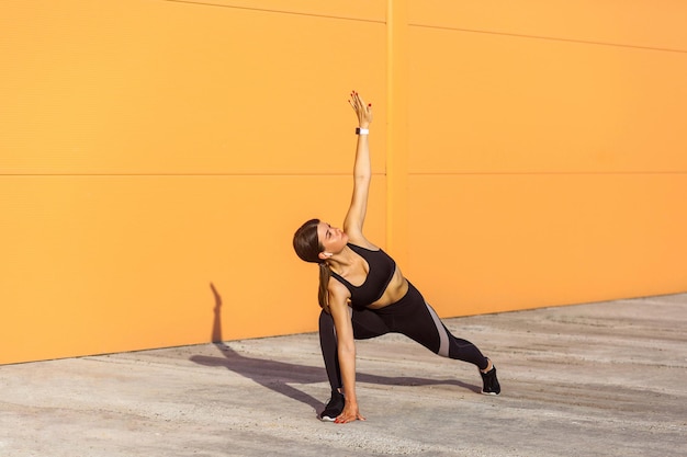 Photo young beautiful yogi woman practicing yoga doing utthita trikonasana exercise extended triangle pose working out wearing sportswear black pants and top outdoor full length orange background