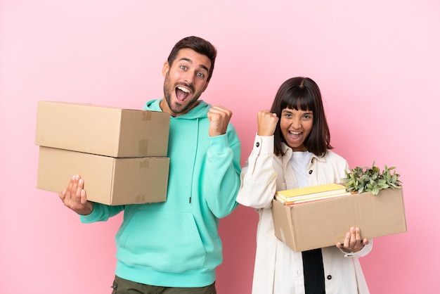 Young beauty couple moving in new home among boxes isolated on pink background celebrating a victory