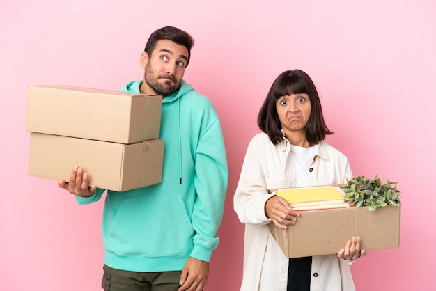 Young beauty couple moving in new home among boxes isolated on pink background making doubts gesture while lifting the shoulders