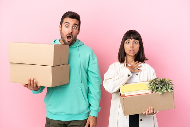 Young beauty couple moving in new home among boxes isolated on pink background with surprise and shocked facial expression